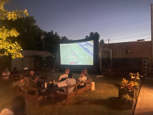people are sitting outside watching a football game on the screen